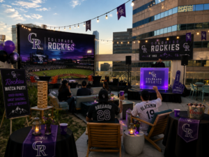 A rooftop Colorado Rockies watch party setup at sunset featuring a giant outdoor projection screen, Rockies-branded banners and decor, string lights, lounge seating, and fans in team jerseys gathered around watching the game against a downtown city skyline backdrop.